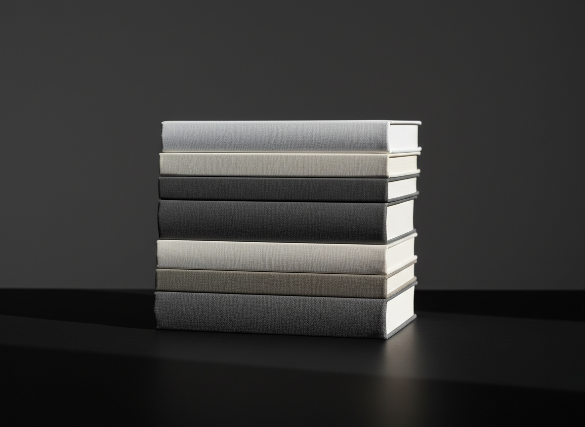 A sleek, minimalist stack of textured hardback books in varying shades of white and charcoal, arranged in a perfectly aligned column atop a crisp matte black desk. The background is a smooth, unfussy deep gray, enhancing the clarity of the composition. Dramatic side lighting from the right casts sharp, angular shadows across the scene, creating high-contrast highlights along the book spines. Shot at a slightly elevated, eye-level perspective with centered composition and sharp focus throughout, the overall atmosphere is bold, modern, and purposeful. The photographic realism and minimalist aesthetic underscore a sense of academic seriousness and professional polish, perfectly reflecting the tone of a teaching portfolio.